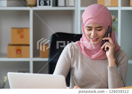 Muslim woman managing online sales, taking phone calls, and organizing packages in a modern workspace. 126038522