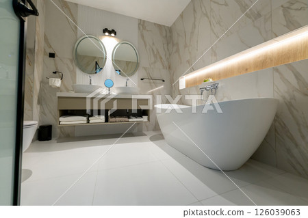 A spacious elegant modern bathroom with freestanding tub, double sink and round mirrors in hotel interior 126039063