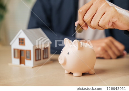 A businessman hand saving money by placing a coin into a piggy bank next to a house model, representing investment, financial stability, property goals, and a savings-focused approach to home buying. A businessman hand saving money by placing a coin into a piggy bank next to a house model, representing investment, financial stability, property goals, and a savings-focused approach to home buying. 126042931