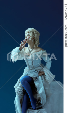 Noble whisper. Young woman in rococo dress with elaborate hairstyle softly gesturing with intrigue against blue background. Noble whisper. Young woman in rococo dress with elaborate hairstyle softly gesturing with intrigue against blue background. 126043741