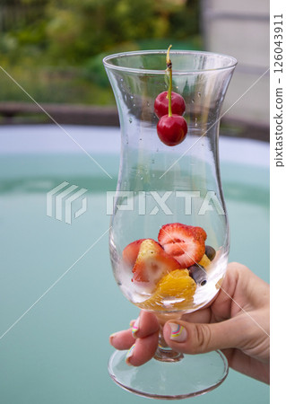 A summer cocktail with strawberries, cherries and foamy champagne in a woman's hand, against a blue pool backdrop. Party concept, vertical 126043911
