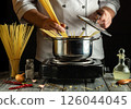A chef stirs spaghetti in a pot on a stovetop, surrounded by fresh ingredients like garlic and chili peppers. The warm lighting creates an inviting atmosphere 126044045