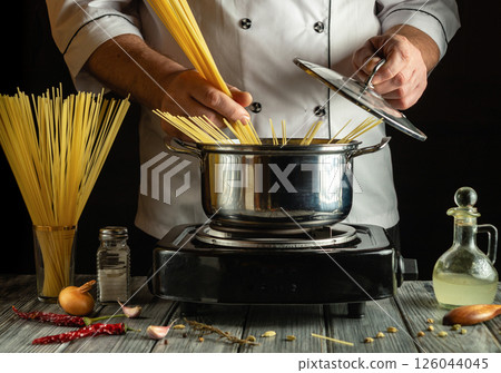 A chef stirs spaghetti in a pot on a stovetop, surrounded by fresh ingredients like garlic and chili peppers. The warm lighting creates an inviting atmosphere 126044045