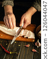 Hands skillfully slice fresh pork belly on a cutting board, surrounded by garlic cloves and dried chilies. The rustic kitchen adds to the authentic cooking atmosphere 126044047