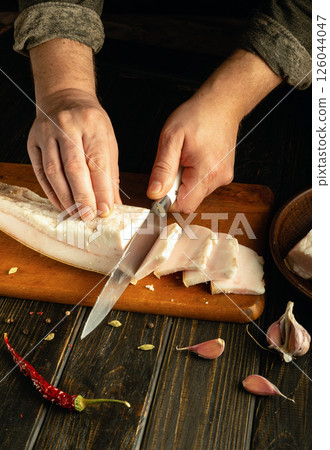 Hands skillfully slice fresh pork belly on a cutting board, surrounded by garlic cloves and dried chilies. The rustic kitchen adds to the authentic cooking atmosphere 126044047