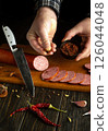 A person slices smoked sausage on a wooden cutting board while holding dried spices in one hand, surrounded by fresh garlic and chili. A sharp knife rests nearby 126044048