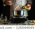 A chef is skillfully dropping sausages into a pot on the stove while holding a green plate with more sausages. Ingredients are scattered on the kitchen counter 126044049