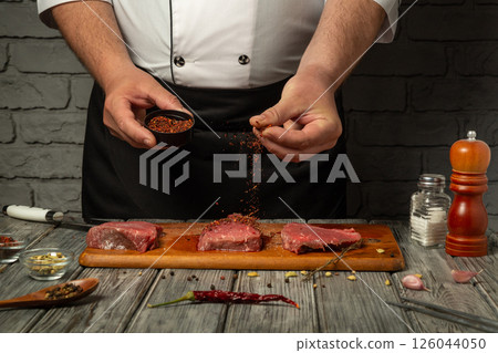A chef sprinkles spices over fresh cuts of beef on a wooden board. The rustic kitchen ambiance enhances the focus on the cooking process, emphasizing culinary skills and flavors A chef sprinkles spices over fresh cuts of beef on a wooden board. The rustic kitchen ambiance enhances the focus on the cooking process, emphasizing culinary skills and flavors 126044050