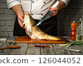 A skilled chef expertly fillets a freshly caught fish on a wooden cutting board. Various ingredients and kitchen tools are arranged nearby, enhancing the culinary atmosphere 126044052