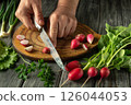 Hands carefully slice crisp radishes on a wooden board surrounded by fresh greens and herbs, showcasing a vibrant home cooking activity 126044053