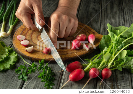 Hands carefully slice crisp radishes on a wooden board surrounded by fresh greens and herbs, showcasing a vibrant home cooking activity Hands carefully slice crisp radishes on a wooden board surrounded by fresh greens and herbs, showcasing a vibrant home cooking activity 126044053
