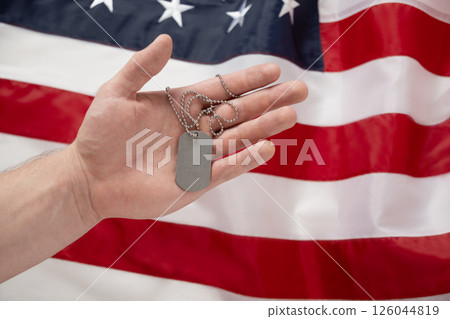 Mans hand holds military soldiers tag against backdrop of American flag. National pride and military tribute with focus on tag. Memorial Day. 126044819