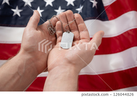 Mans hands holds military dog tag against backdrop of American flag. National pride and military tribute with focus on tag. Memorial Day. 126044826