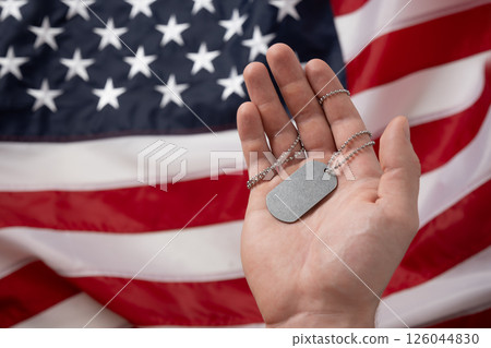 Mans hand holds military soldiers tag against backdrop of American flag. National pride and military tribute with focus on tag. Memorial Day. Mans hand holds military soldiers tag against backdrop of American flag. National pride and military tribute with focus on tag. Memorial Day. 126044830