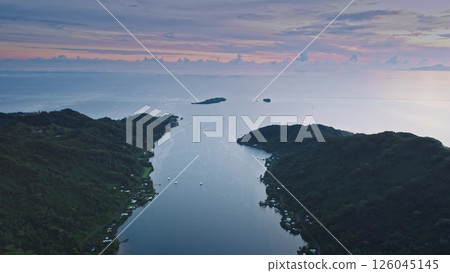 Tranquil aerial view captures a serene coastal landscape. The scene features lush green hills and a calm bay, set against a vibrant sunset sky with scattered clouds. Drone fly over sea ocean water 126045145