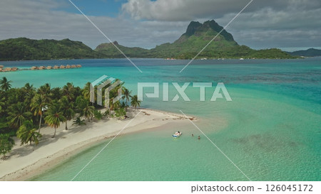 Crystal-clear turquoise water gently laps the shore of a pristine white sand beach nestled beside lush palm trees, with tourists enjoying the breathtaking view of mount Otemanu in Bora Bora 126045172
