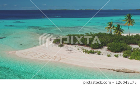 Clear turquoise waters surround a sandy beach where 'HOLIDAY' is written in the sand. Lush greenery contrasts against the vibrant sea, creating a picturesque tropical setting. 126045173