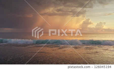 Golden hour sunlight bathing ocean waves as they crash onto the shore, while distant dark clouds release rain, creating a dramatic and scenic seascape 126045194