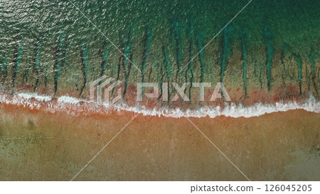 An aerial perspective showcases a calm shoreline where gentle waves meet the sandy beach. The interplay of water and land creates a tranquil scene during sunset hours. 126045205