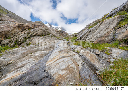 A stunning view of the Schlaten Glacier area in Austria, featuring rugged, layered rocky terrain illuminated by soft sunlight under a partly cloudy sky. The landscape showcases the beauty of Alps 126045471