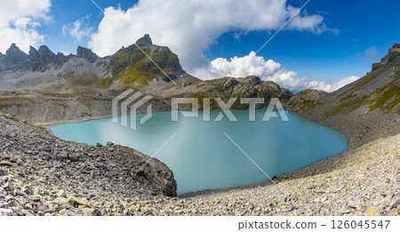 Turquoise alpine lake Wildsee surrounded by rocky mountain peaks and grass-covered slopes under a partly cloudy sky in the Pizol region. The natural beauty of the serene landscape is highlighted 126045547