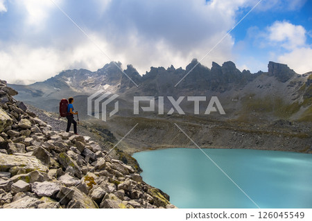Hiker with a red backpack and poles admires the view of Pizol Lake Wildsee in a breathtaking mountain landscape. The turquoise water, rocky peaks. Travel sport wanderlust concept. 126045549