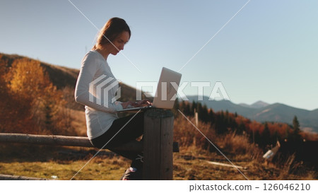 Woman typing on laptop and enjoying autumn sunset mountains nature. Work outdoor, online digital nomad remote job in Carpathian mountains. Outdoor lifestyle travel on summer holiday vacation. 126046210