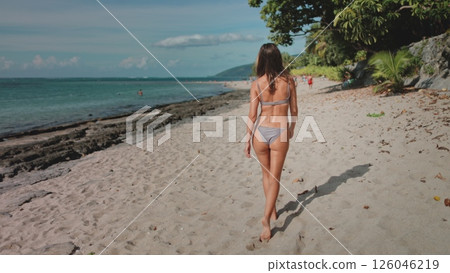 A stunning woman wearing a bikini is leisurely walking down the sandy beach on a beautiful summer day. The sun is gently setting in the background, casting a warm glow over the scene. 126046219