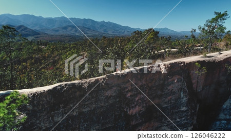 Aerial drone footage of Pai Canyon in northern Thailand shows a tourist walking along a narrow path on a cliff overlooking a valley with mountains in the background 126046222