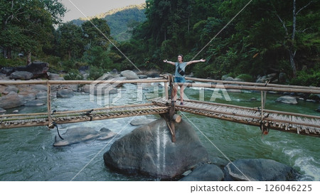Young woman tourist is standing with open arms on a bamboo bridge over a wild river flowing through a tropical forest, enjoying the freedom and beauty of nature 126046225