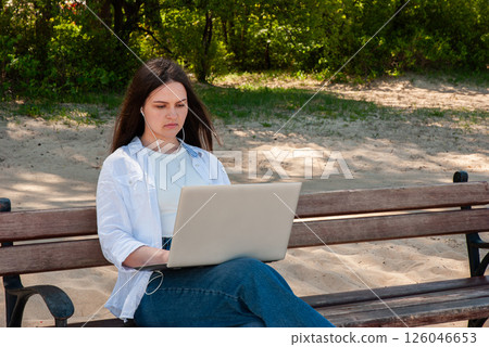 Young caucasian female using laptop with earphones on park bench on sunny day, focused on work and enjoying nature's tranquility. Digital nomad lifestyle. Remote work 126046653