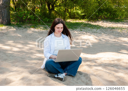 Caucasian young adult female in casual attire focuses on laptop while seated on a sunny sandy beach under the shade of lush green trees. Digital nomad lifestyle. Remote work Caucasian young adult female in casual attire focuses on laptop while seated on a sunny sandy beach under the shade of lush green trees. Digital nomad lifestyle. Remote work 126046655