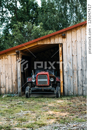Old red tractor parked inside a wooden shed surrounded by trees in a rural setting. Agricultural heritage, rural economy, self-sufficiency, traditional farming.. 126047417