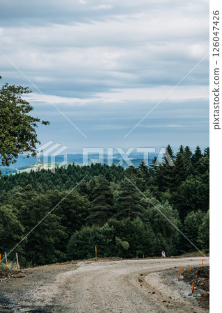 Dirt road under construction winding through a dense forested landscape with mountains in the background. Infrastructure expansion, forest development, land modification, remote access routes.. 126047426