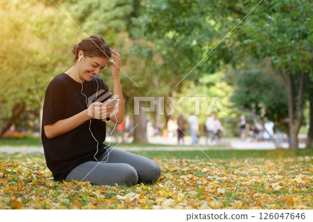 Young beautiful girl sits in park, uses smartphone, listens to music and laughs 126047646