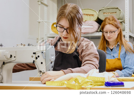 Two female seamstresses in the workshop at the factory are working at tables on sewing machines.  126048698