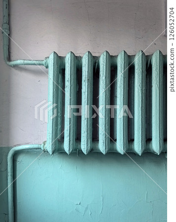 An old central heating radiator in the entrance of an apartment building. The radiator is blue-green in cracked paint against a plaster wall. Heating of buildings during the cold winter season An old central heating radiator in the entrance of an apartment building. The radiator is blue-green in cracked paint against a plaster wall. Heating of buildings during the cold winter season 126052704