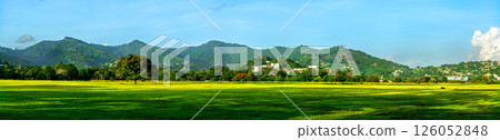 A wide panoramic view of Queen's Park Savannah in Port of Spain, Trinidad, featuring a vast green field bordered by trees and backed by the lush Northern Range mountains under a clear sky. 126052848