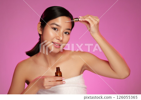 Woman applying skincare serum in a bright studio with pink background during a beauty routine 126055365