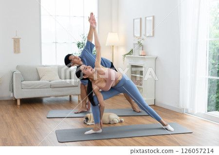 Couple engages in yoga practice on mats in a well-lit living room. A small dog rests nearby, while the sunlight illuminates the serene space, creating a calm atmosphere. 126057214