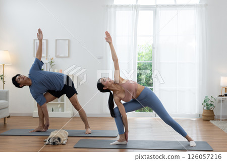 Couple engages in yoga practice on mats in a well-lit living room. A small dog rests nearby, while the sunlight illuminates the serene space, creating a calm atmosphere. Couple engages in yoga practice on mats in a well-lit living room. A small dog rests nearby, while the sunlight illuminates the serene space, creating a calm atmosphere. 126057216