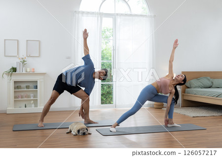 Couple engages in yoga practice on mats in a well-lit living room. A small dog rests nearby, while the sunlight illuminates the serene space, creating a calm atmosphere. Couple engages in yoga practice on mats in a well-lit living room. A small dog rests nearby, while the sunlight illuminates the serene space, creating a calm atmosphere. 126057217