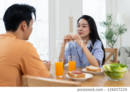 Couple enjoying breakfast are seated at wooden table in a bright kitchen, sharing breakfast. They have orange juice, salad, and pastries while engaging in cheerful conversation. Couple enjoying breakfast are seated at wooden table in a bright kitchen, sharing breakfast. They have orange juice, salad, and pastries while engaging in cheerful conversation. 126057229