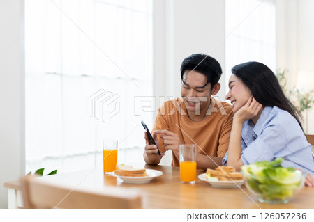 Couple enjoying together while laughing look at smartphone in dining table with glasses of orange juice and breakfast items are present in bright bedroom background. 126057236