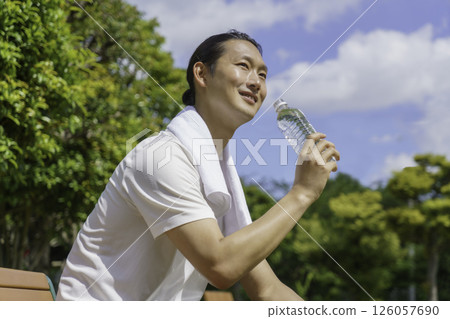 一位年輕人坐在公園長椅上補充水分 126057690