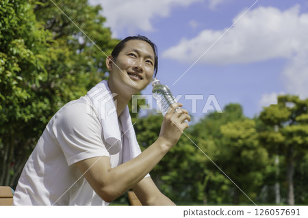 一位年輕人坐在公園長椅上補充水分 126057691