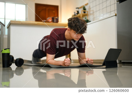 Young man doing plank exercise at home while following a workout video on a tablet 126060600