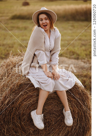 Young woman in beige dress sitting on haystack in village field 126060710