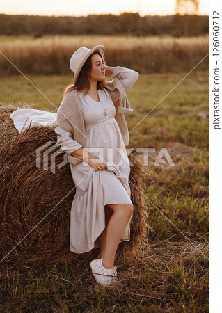 A woman stands in a summer field at golden hour leaning on a bale of hay. 126060712