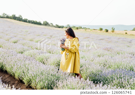 woman in yellow dress stands in lavender field, gently holding bouquet of lavender flowers. She appears serene and contemplative, enjoying beauty and fragrance of blossoms around her. woman in yellow dress stands in lavender field, gently holding bouquet of lavender flowers. She appears serene and contemplative, enjoying beauty and fragrance of blossoms around her. 126065741